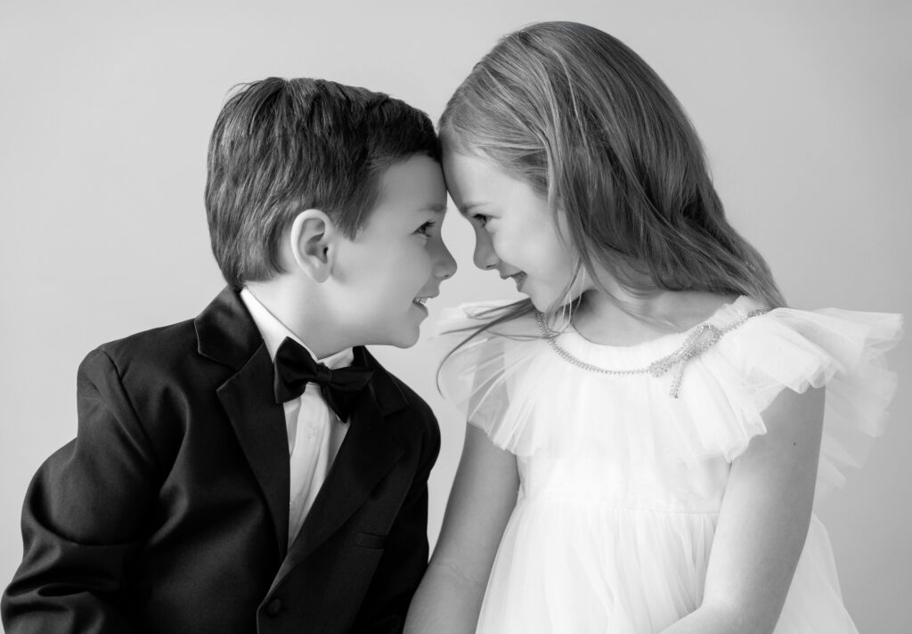 Black and white photo of two siblings, forehead to forehead, dressed up in formal wear.