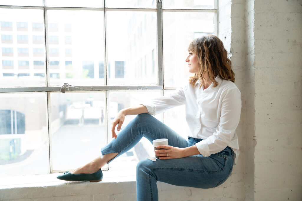 woman sitting in window sill looking out holding a coffee mug
